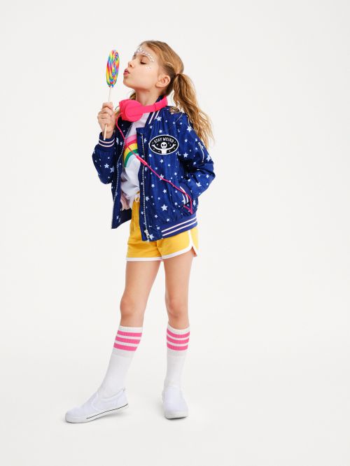 Young girl in blue star jacket poses with colorful lollipop. She wears yellow shorts, striped socks, and pink headphones in studio shot.
