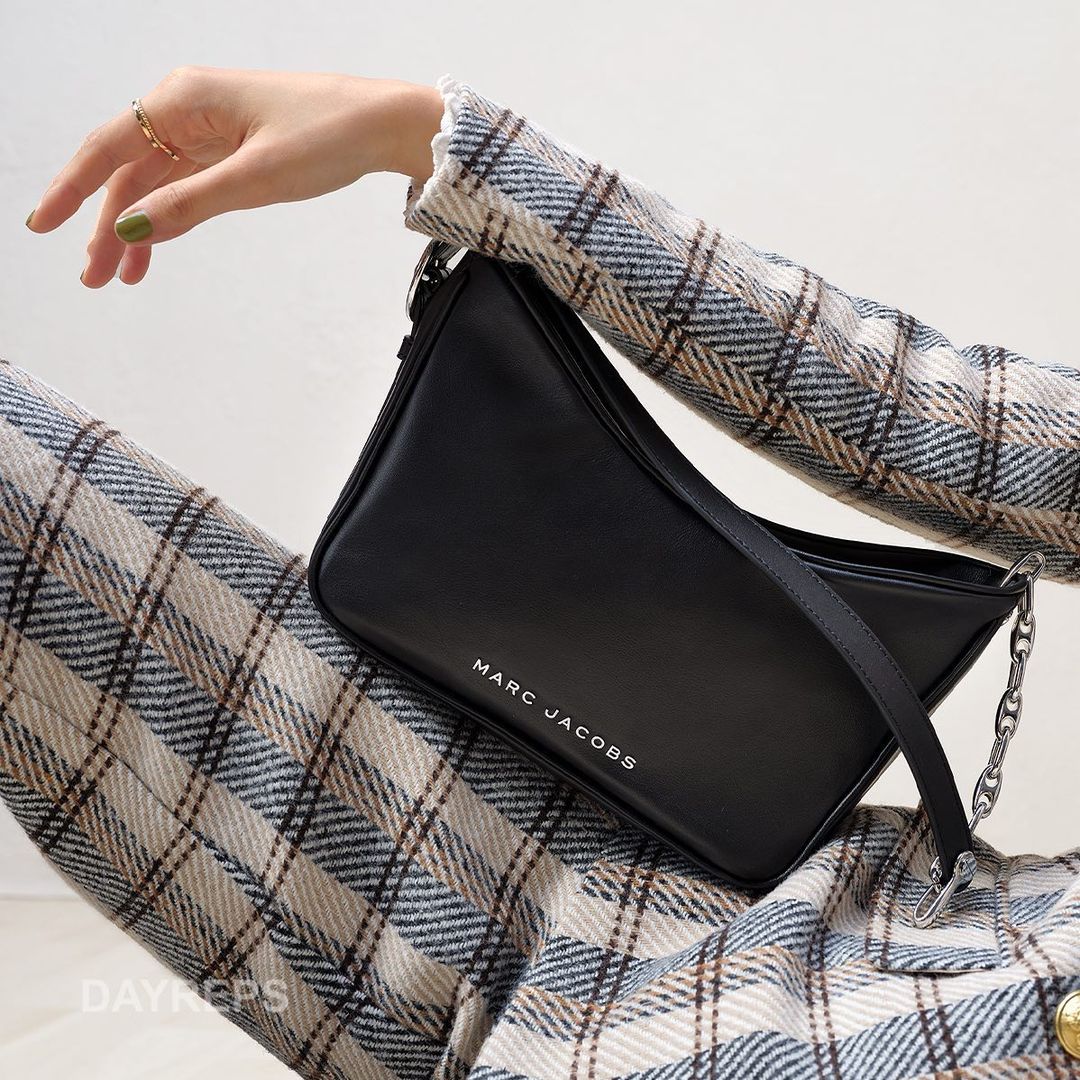 Black Marc Jacobs handbag on a plaid background with a hand wearing a gold ring and green nail polish.