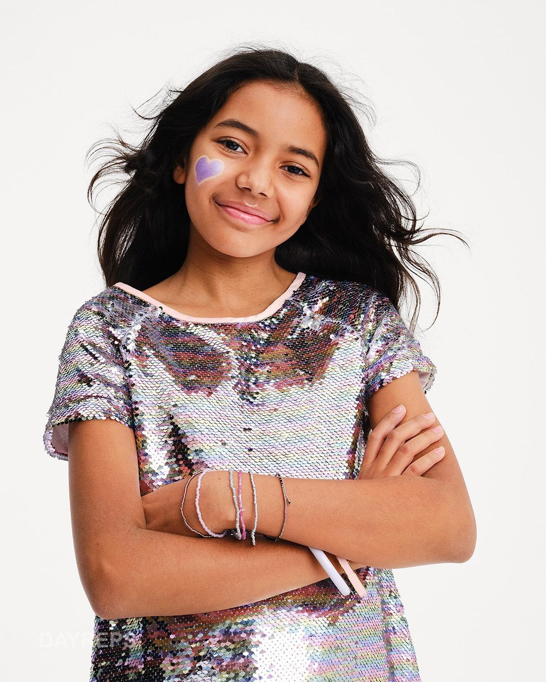 Young girl with heart face paint smiles in a sequin top against a bright backdrop, creating a joyful and engaging portrait.