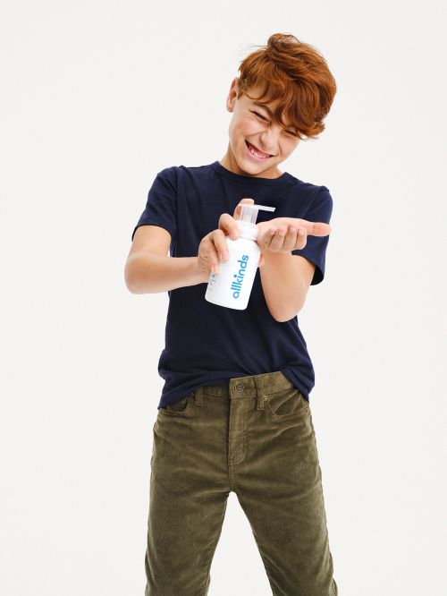 Red-haired child applies liquid soap from a white bottle with a pump to the palm of their hand in a brightly lit indoor setting.