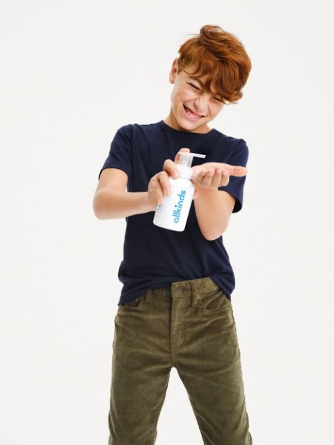 Red-haired child applies liquid soap from a white bottle with a pump to the palm of their hand in a brightly lit indoor setting.