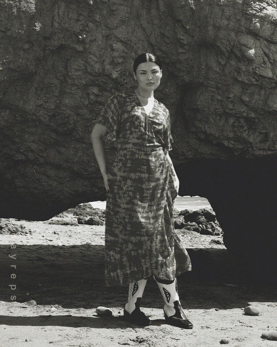 Woman in patterned dress and cowboy boots poses in front of a large rock formation on a sandy beach in a black and white photo.