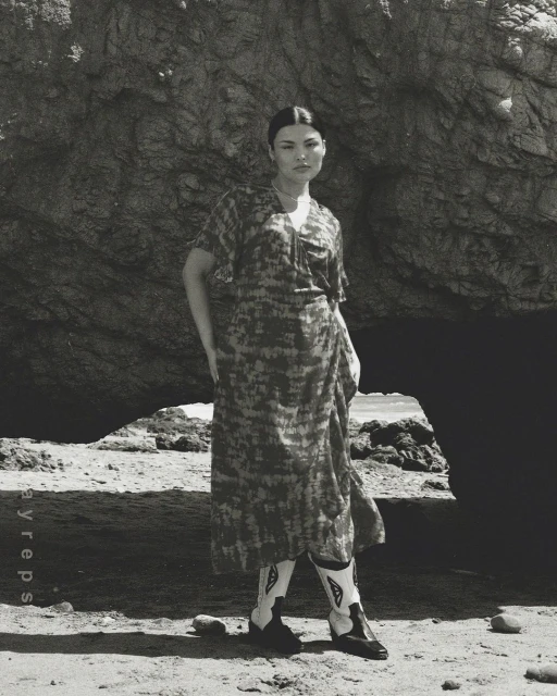 Woman in patterned dress and cowboy boots poses in front of a large rock formation on a sandy beach in a black and white photo.