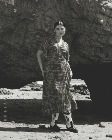 Woman in patterned dress and cowboy boots poses in front of a large rock formation on a sandy beach in a black and white photo.