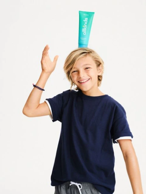 A young boy with blonde hair smiles while balancing a tube of Allkinds skincare on his head, wearing a navy blue t-shirt indoors.
