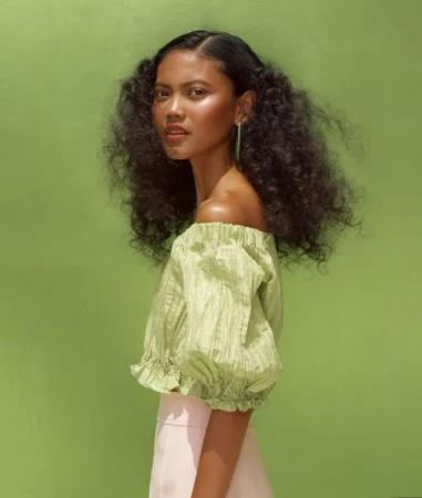 Woman with dark curly hair in off-shoulder green top and pink skirt poses in front of a green background. Beauty and fashion concept.