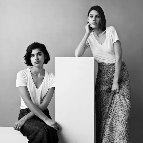 Two adult females pose in a monochromatic photo studio. One sits, another leans against a white box in casual fashion attire.