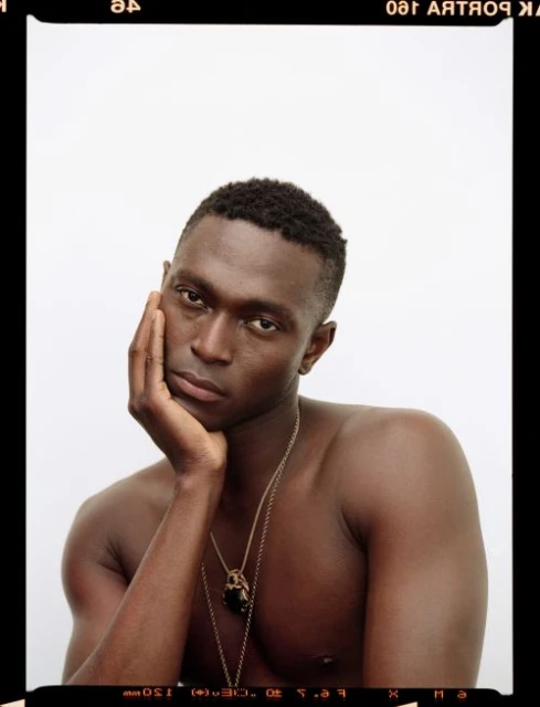Striking portrait of a Black man with bare chest, hand on face, wearing pendant necklace against a bright white studio backdrop.