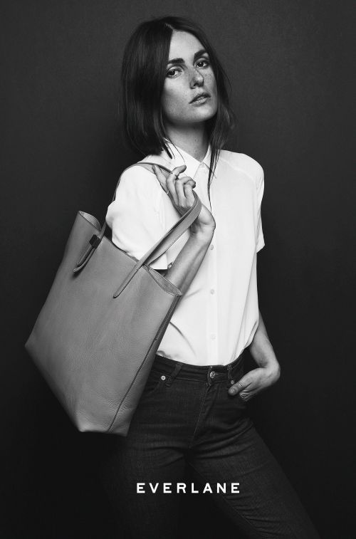 Indoor portrait of a woman with medium-length hair holding a tan tote bag while wearing a white shirt and dark jeans in a photo studio.