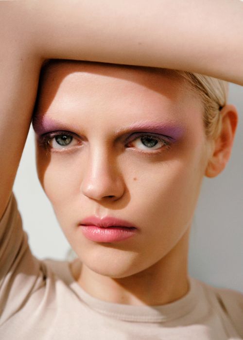 Close-up portrait of a woman with purple eyeshadow and pink lipstick posing with her arm raised above her head.