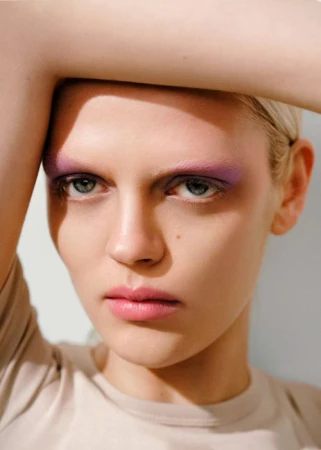 Close-up portrait of a woman with purple eyeshadow and pink lipstick posing with her arm raised above her head.