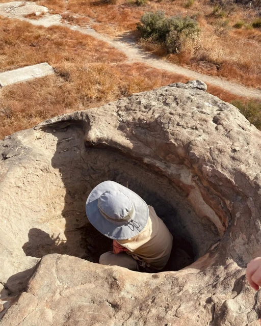 Person wearing a hat crouched inside a large rock formation in a dry, grassy landscape, possibly exploring or examining the rock.