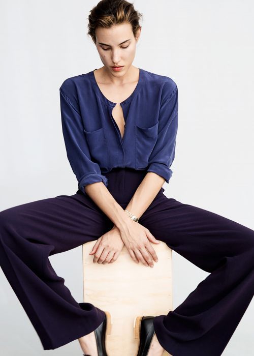 Model wearing a blue buttoned blouse and dark trousers seated on a wooden box against a white background
