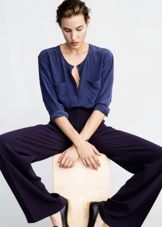 Model wearing a blue buttoned blouse and dark trousers seated on a wooden box against a white background