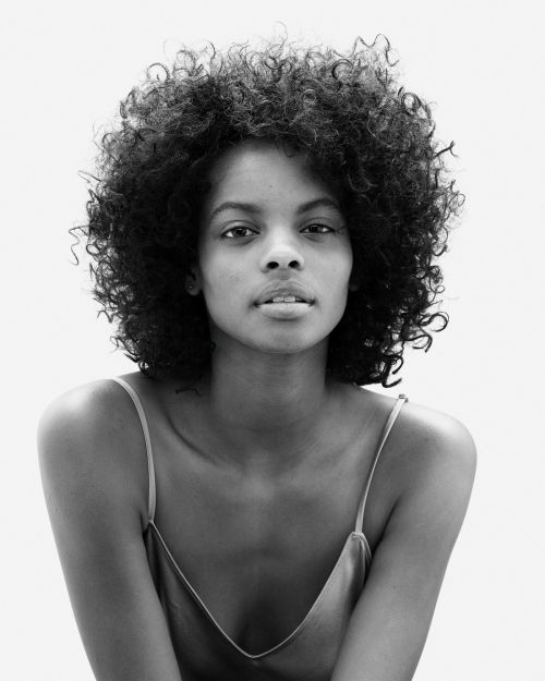 Black and white portrait of a woman with curly hair wearing a camisole top against a white background in eye-level, medium shot.