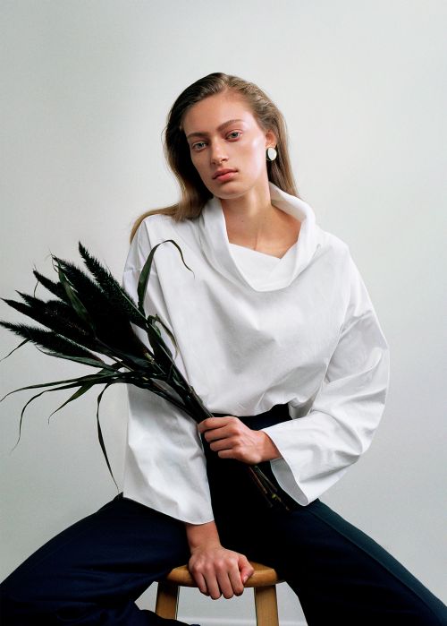 Woman in a white top poses indoors while seated and holding dark foliage. She has long hair and sits in front of a bright white backdrop.