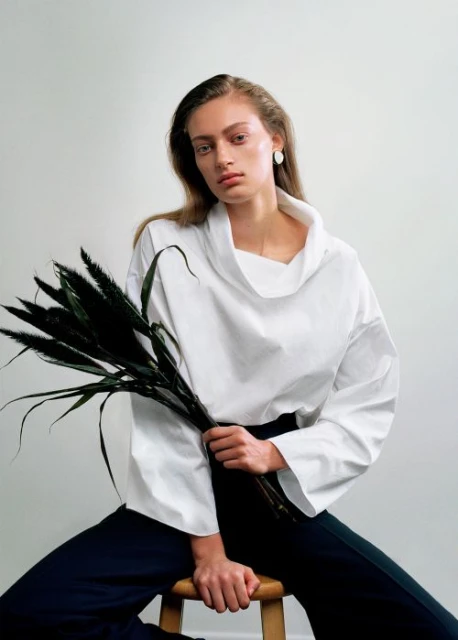 Woman in a white top poses indoors while seated and holding dark foliage. She has long hair and sits in front of a bright white backdrop.