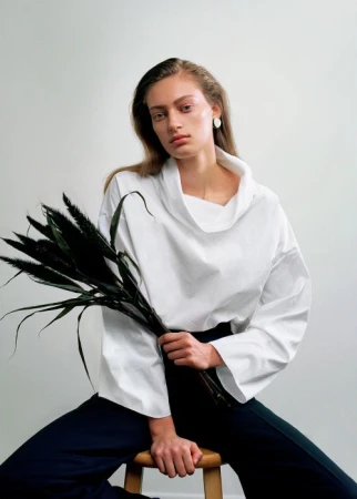 Woman in a white top poses indoors while seated and holding dark foliage. She has long hair and sits in front of a bright white backdrop.