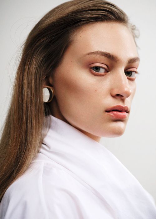 Close-up portrait of a young adult woman with long brown hair, wearing a white collared shirt and statement earrings against a white background.
