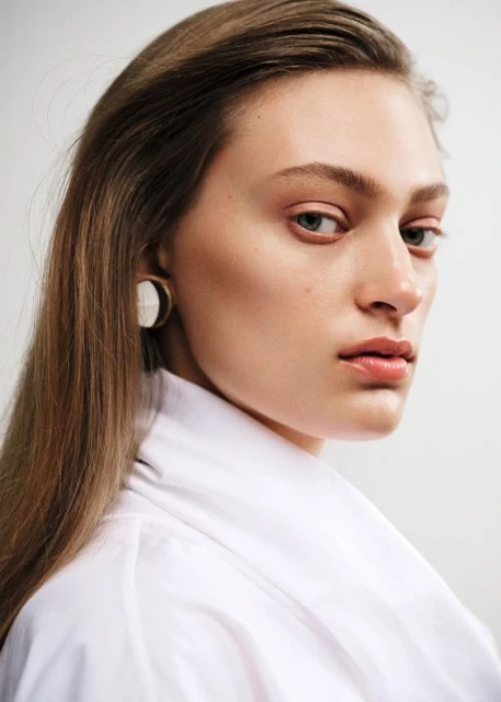 Close-up portrait of a young adult woman with long brown hair, wearing a white collared shirt and statement earrings against a white background.
