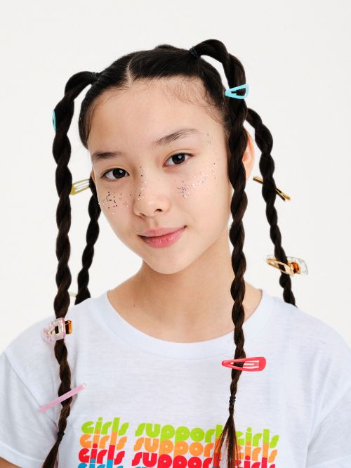 Portrait of a young girl with twisted pigtails adorned with colorful hair clips, wearing a 'Girls Support Girls' t-shirt against a white backdrop.