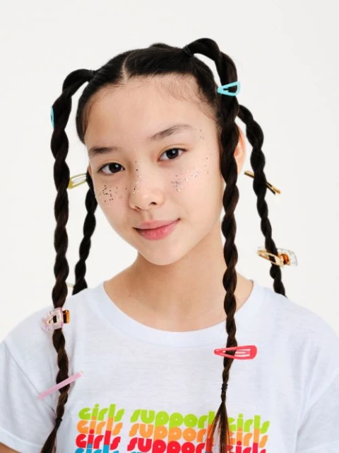 Portrait of a young girl with twisted pigtails adorned with colorful hair clips, wearing a 'Girls Support Girls' t-shirt against a white backdrop.