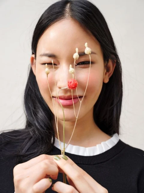 Portrait of a young Asian woman holding dried flowers with a red bud, close to her face, wearing a black sweater with a white collar.