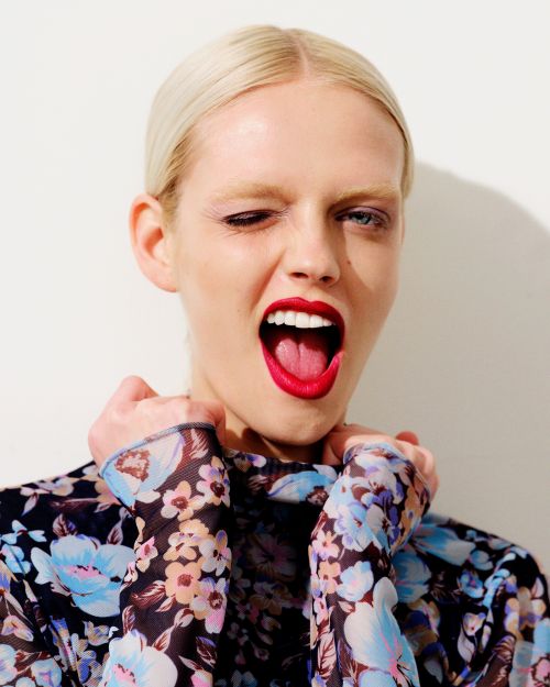 Close-up portrait of a blonde woman wearing a blue floral sheer top, winking and laughing with bright red lipstick by Kathryna Hancock.