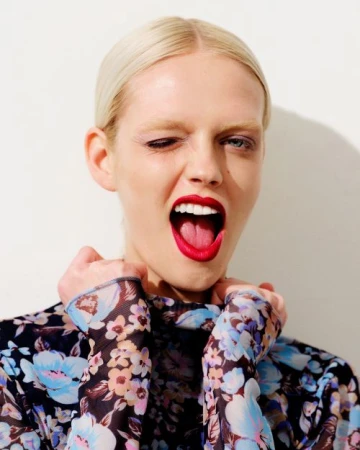 Close-up portrait of a blonde woman wearing a blue floral sheer top, winking and laughing with bright red lipstick by Kathryna Hancock.
