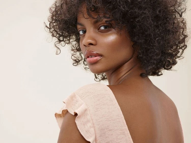 Close-up portrait of a young Black woman with curly hair looking over her shoulder wearing a peach-colored top in a studio setting.