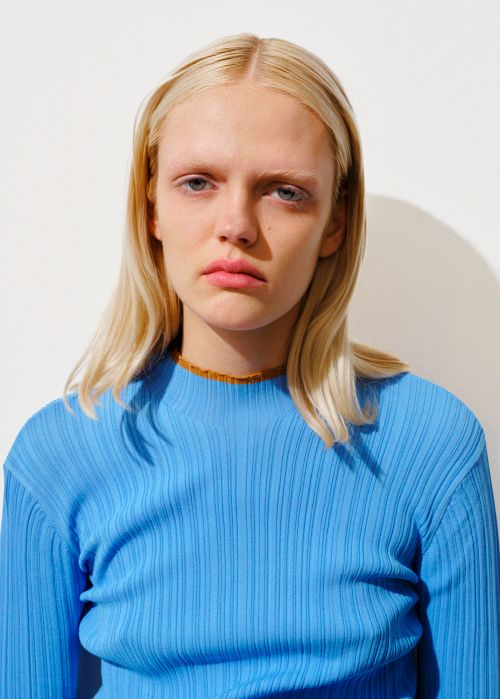 Portrait of a blonde woman wearing a vibrant blue ribbed top against a plain white wall with visible shadow.