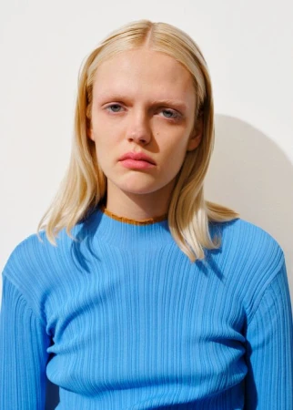 Portrait of a blonde woman wearing a vibrant blue ribbed top against a plain white wall with visible shadow.