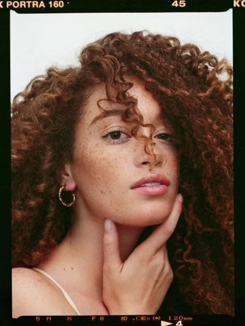 Close-up portrait of a beautiful woman with curly auburn hair and freckles, her hand gently touching her face, studio lighting.