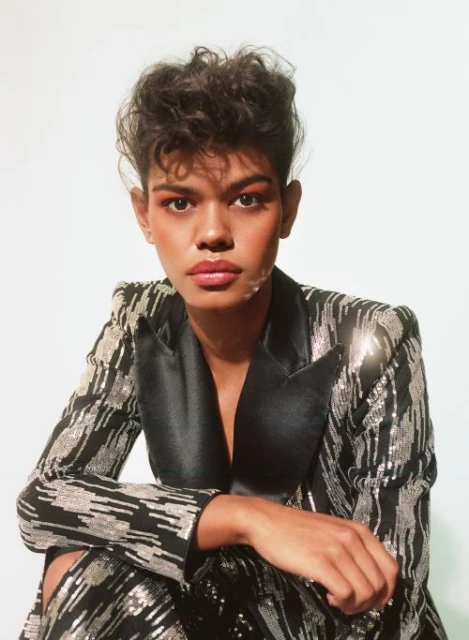 Close-up portrait of a person with curly dark hair wearing a black blazer with silver embellishments against a neutral backdrop.