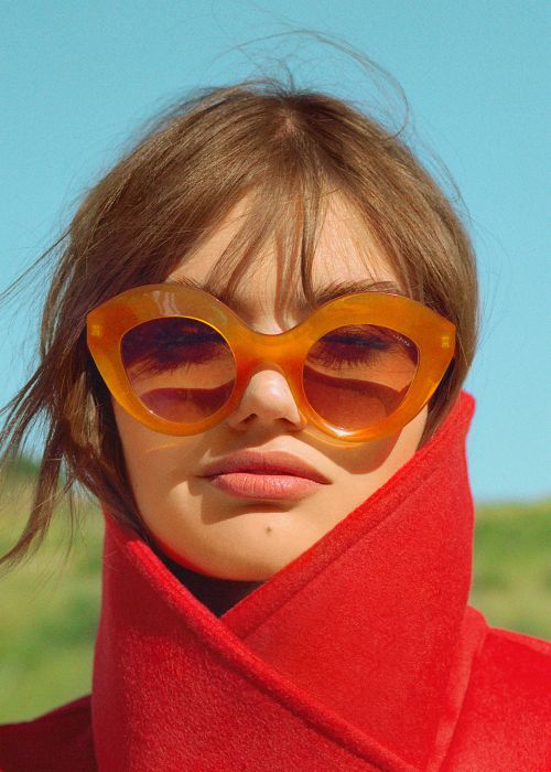 Headshot of a woman with orange sunglasses and a red coat against a background with a blue sky and green grass in natural light.