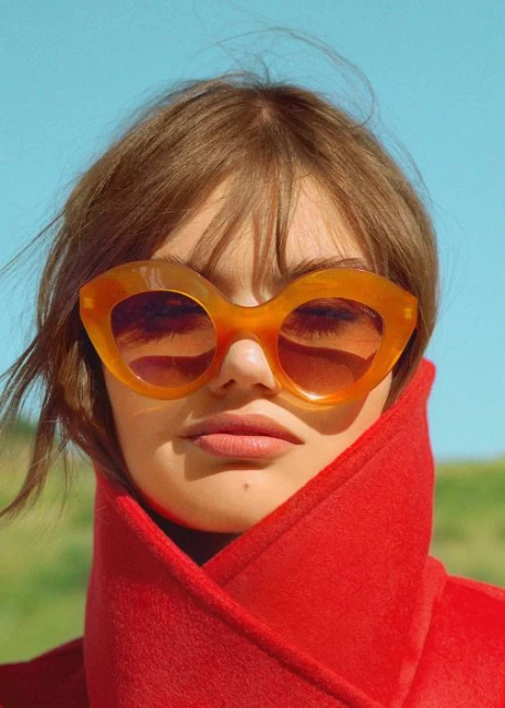 Headshot of a woman with orange sunglasses and a red coat against a background with a blue sky and green grass in natural light.