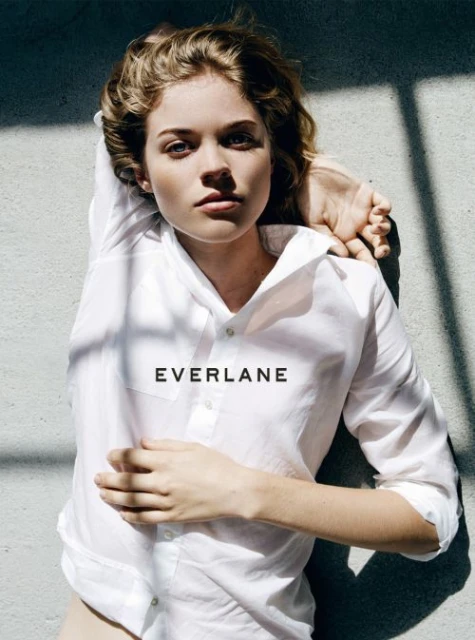 Blonde woman in a white shirt lying on a gray surface with a shadow, staring at the camera. Features the Everlane logo.