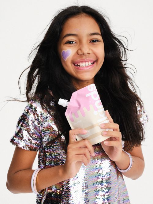 A smiling young girl holds a pouch with a purple heart on her cheek, wearing a sequin top in a white studio setting, portrait shot.