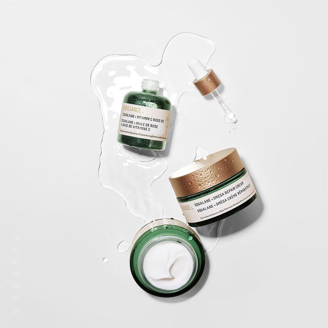 Overhead shot of Biossance Squalane + Vitamin C rose oil and Squalane + Omega repair cream in a minimalistic setting with water droplets.