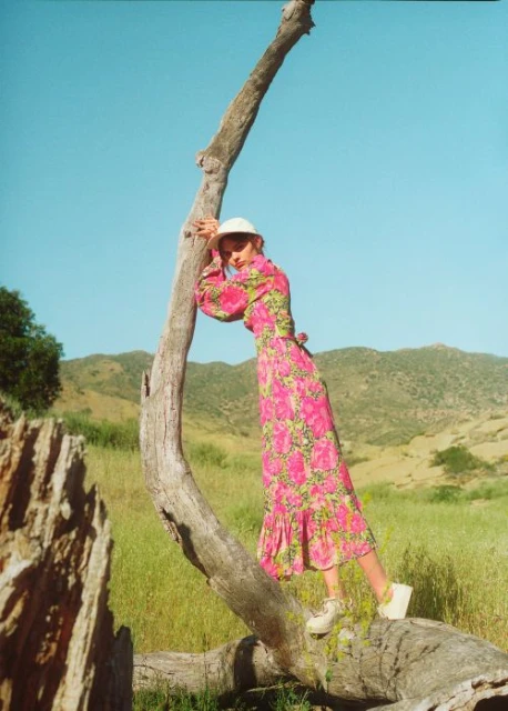 Woman in pink floral dress leans on fallen tree in a grassy field with mountains under a clear sky, posing in outdoor fashion shot.