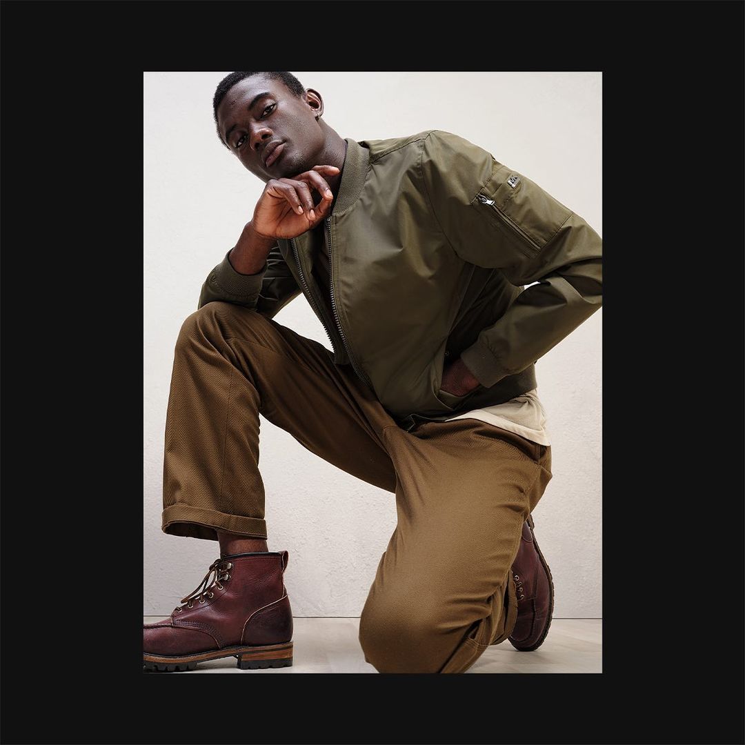 A dark-skinned male model is posed on his knee wearing a green bomber jacket with brown trousers and boots against an off-white background.