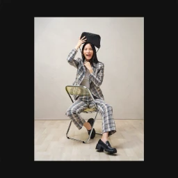 Woman in a plaid pantsuit holding a black handbag above her head while seated on a folding chair in a studio.