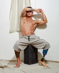 Shirtless man with tattoos and prosthetic leg sitting on dark block against white wall