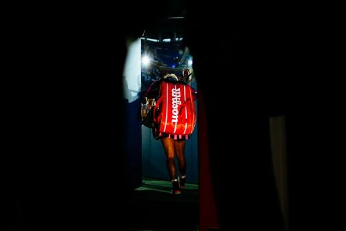 Female tennis player carrying a Wilson branded tennis bag walks from darkness into the light towards the tennis court.
