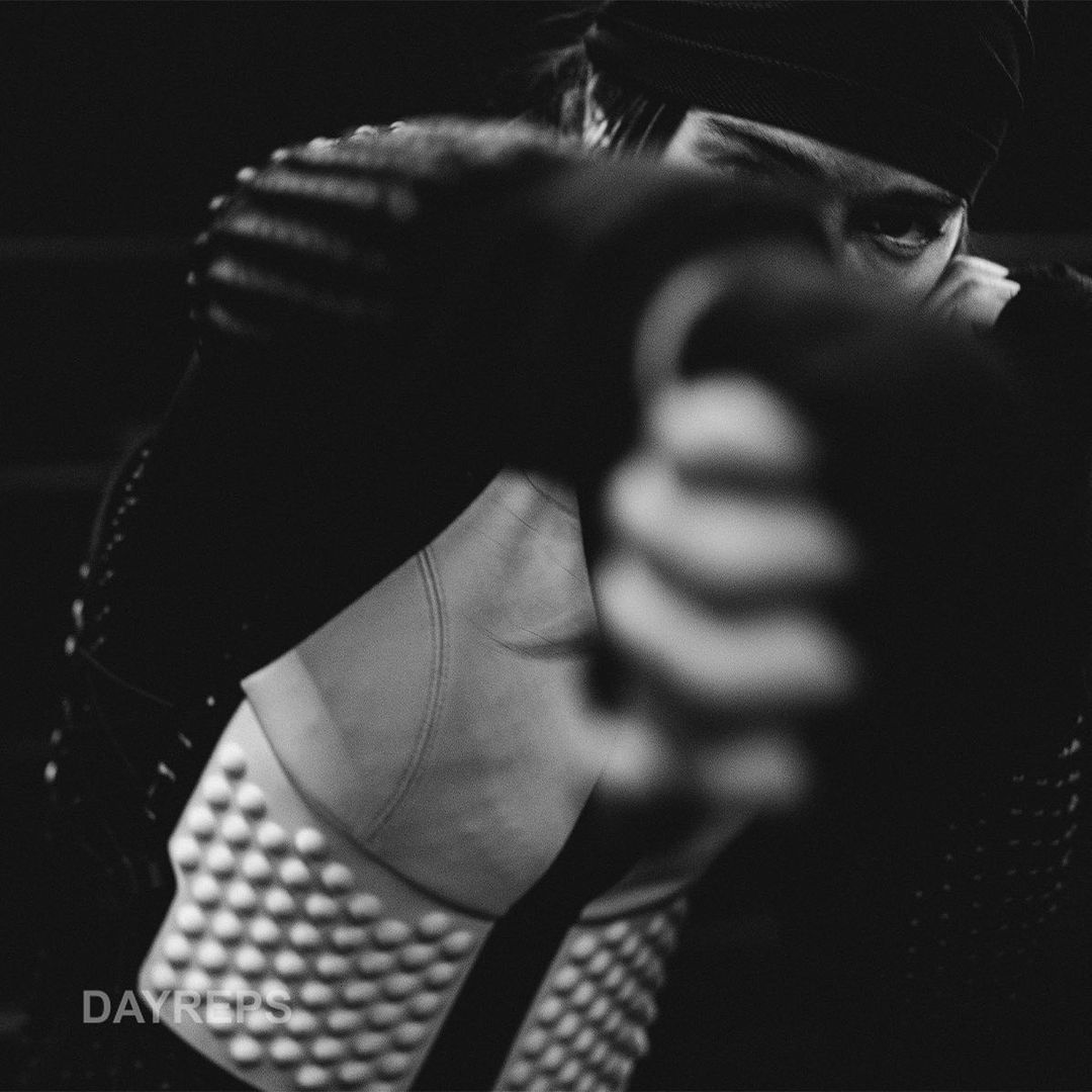 Monochrome portrait of boxer with black headband and studded shoulder pads, fist in foreground, set against a dark background.