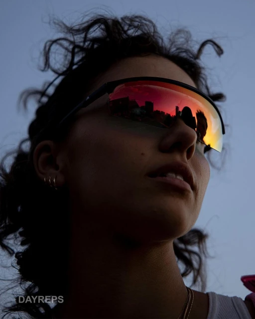 Portrait of a dark-haired woman wearing sporty sunglasses reflecting a cityscape, seen in a low-angle shot against a blue sky.