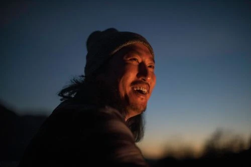 Close-up of Asian adult male wearing a beanie hat and smiling outdoors at night against a dark blue sky.