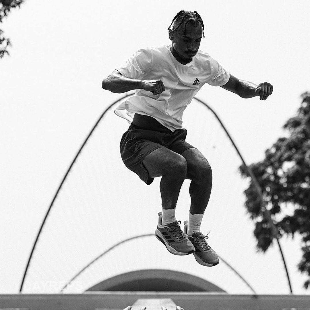 A male athlete in sportswear leaps in the air against an outdoor backdrop with trees and architectural features in a black-and-white photo.
