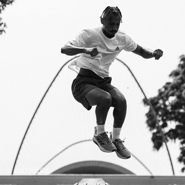 A male athlete in sportswear leaps in the air against an outdoor backdrop with trees and architectural features in a black-and-white photo.