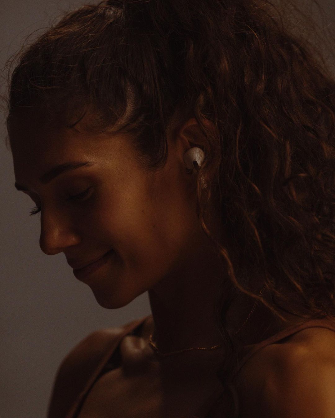 Side view of a smiling adult woman with curly hair and wireless earbuds, softly lit against a neutral studio backdrop.